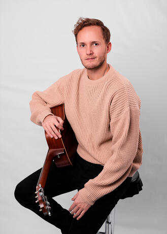 An editorial portrait of a man sitting on a stool in a tan sweater, holding an acoustic guitar.