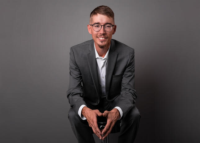A professional headshot of a man wearing a white shirt and grey suit with his hands folded.